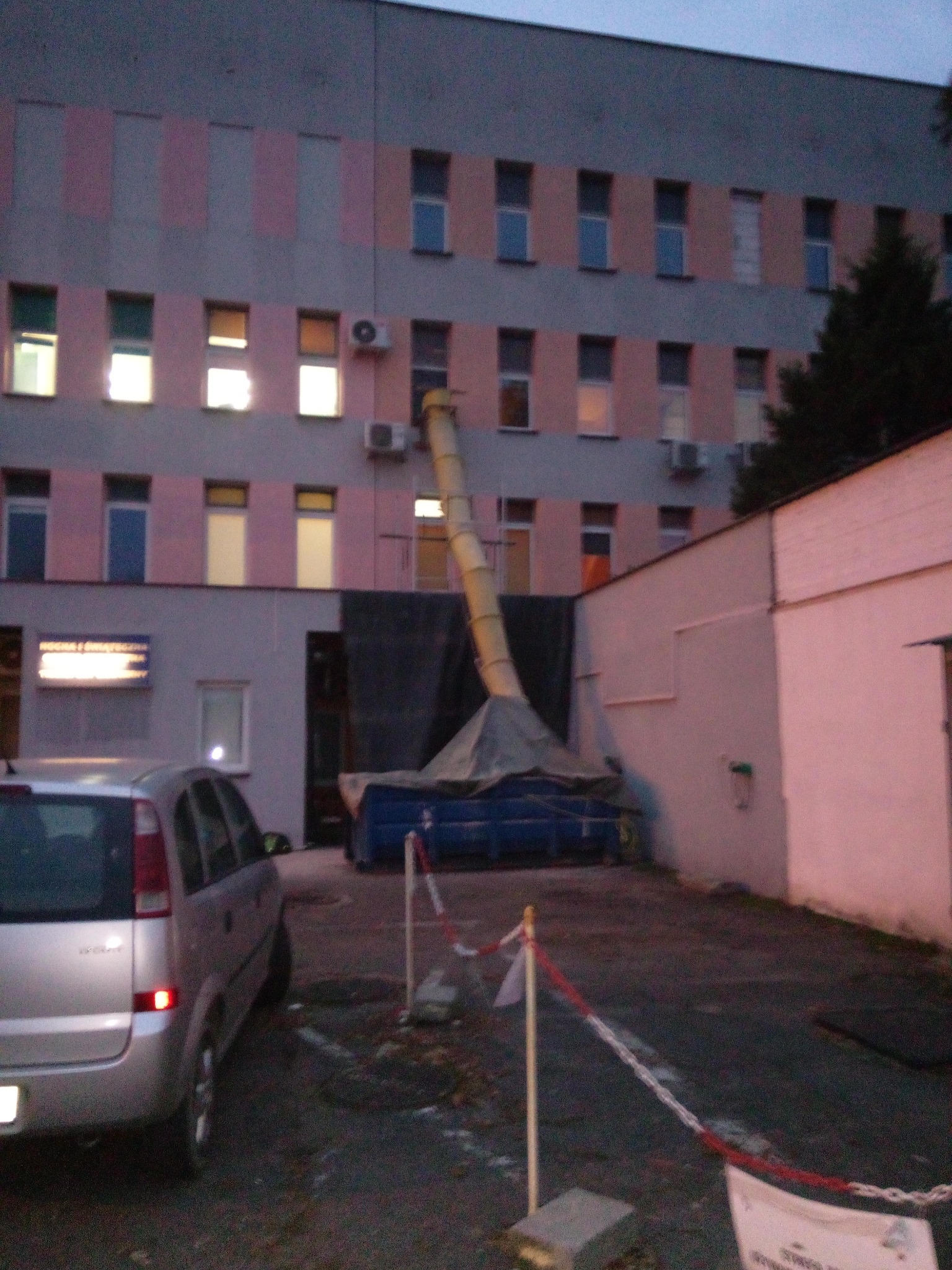 Kontener na gruz podłączony do rury zsypowej na tle budynku z oknami. Widoczny samochód i taśmy ostrzegawcze.