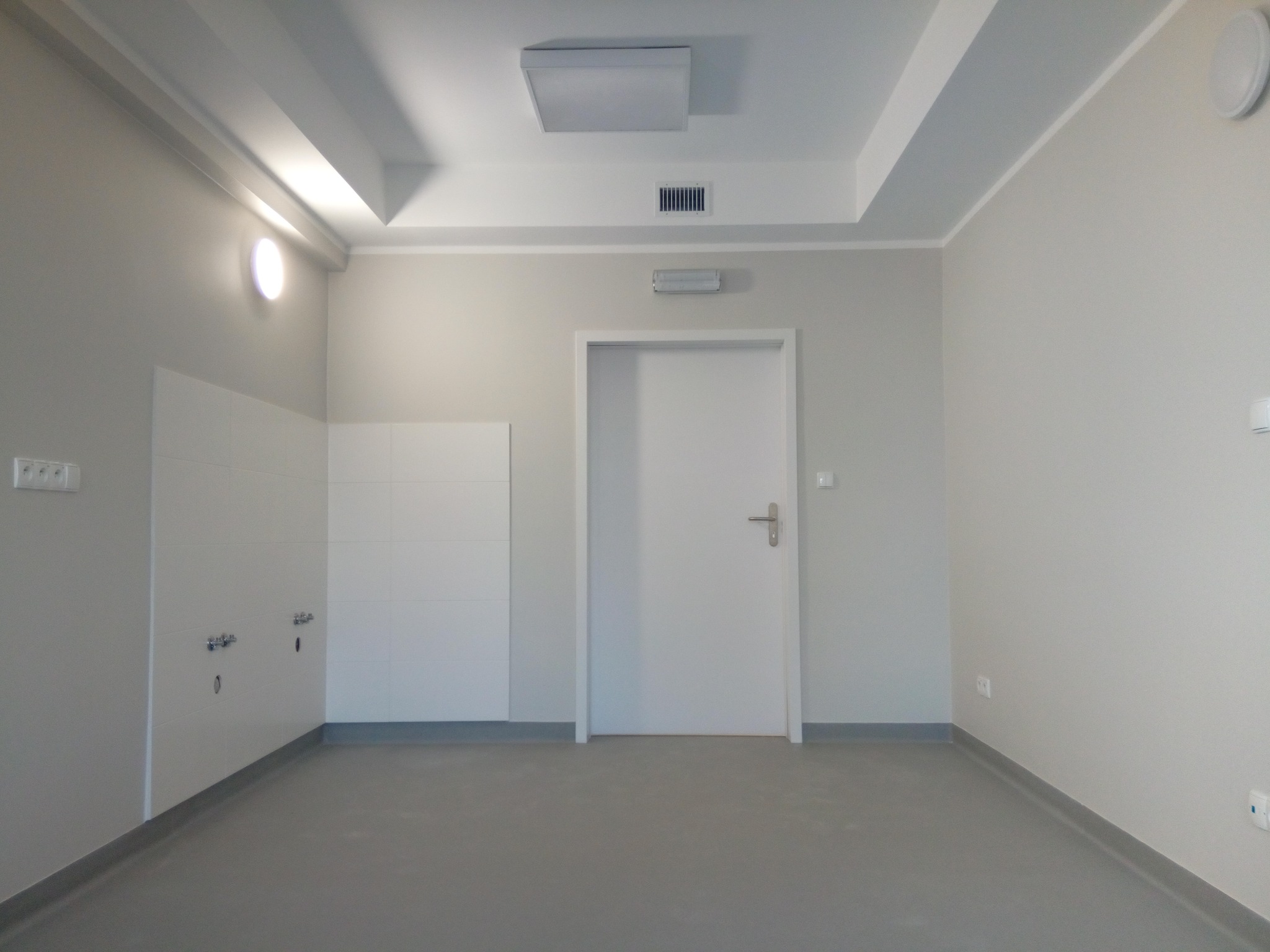 Wnętrze po remoncie: białe ściany, szara podłoga z listwami, białe płytki na ścianie z instalacją wodną, białe drzwi. Minimalistyczny design, neutralna kolorystyka.