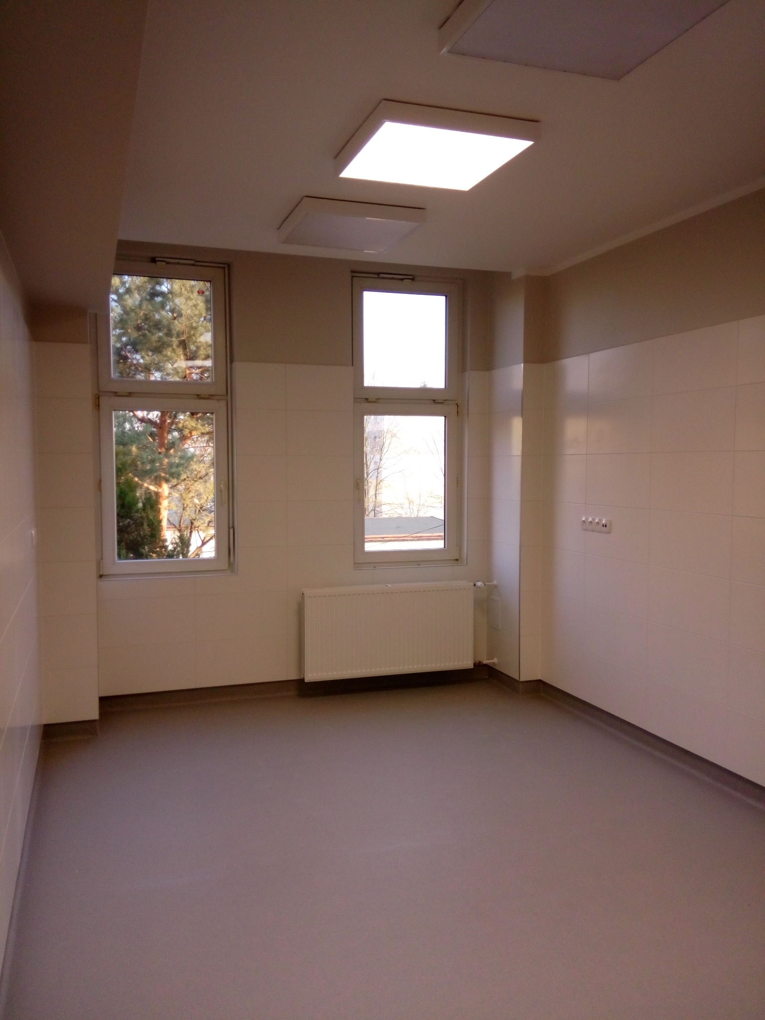 Wnętrze po remoncie: jasne płytki ścienne, szara podłoga, dwa okna z widokiem na drzewa, biały grzejnik i sufitowe lampy. Minimalistyczny, odświeżony pokój.