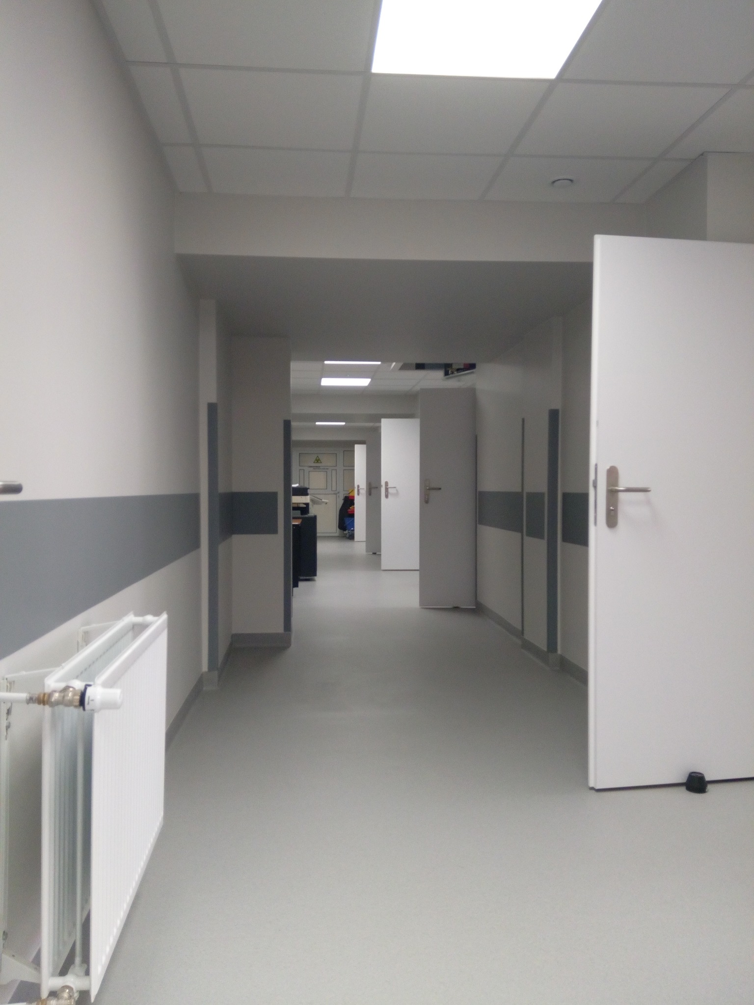 Jasny korytarz w biurze lub placówce medycznej po remoncie, z białymi ścianami, szarymi pasami ochronnymi i grzejnikiem. Widoczne drzwi i oświetlenie sufitowe.
