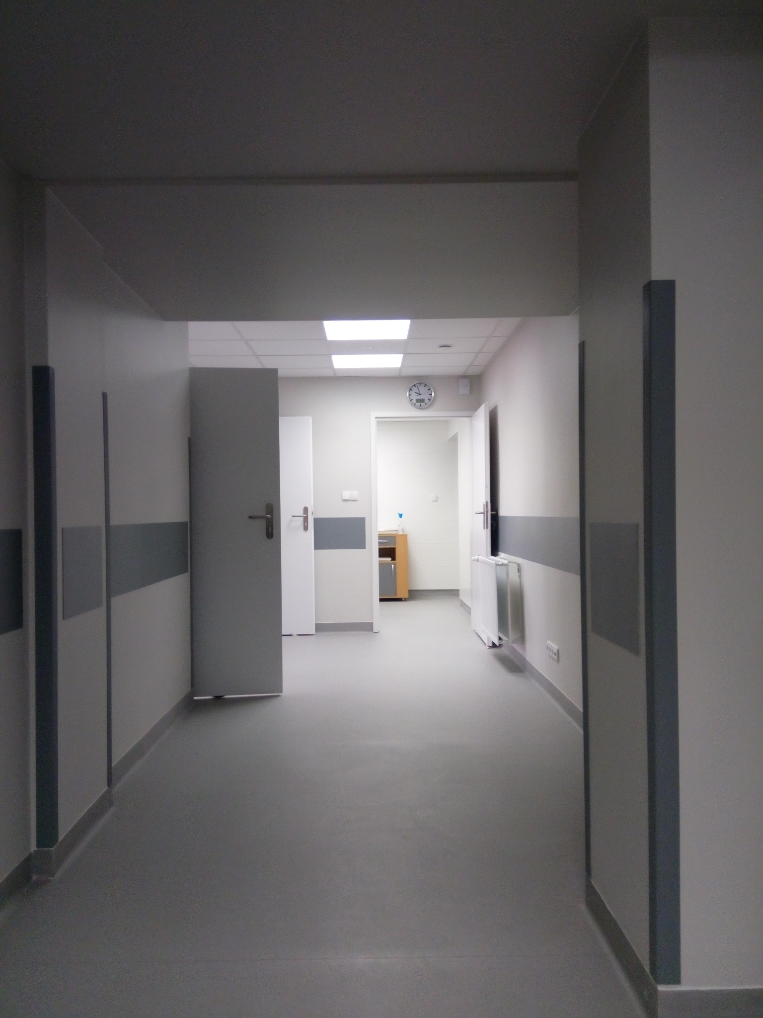 Korytarz w neutralnych barwach z szarymi panelami ściennymi i drzwiami. Widoczny zegar na końcu korytarza, oświetlenie sufitowe i grzejnik.