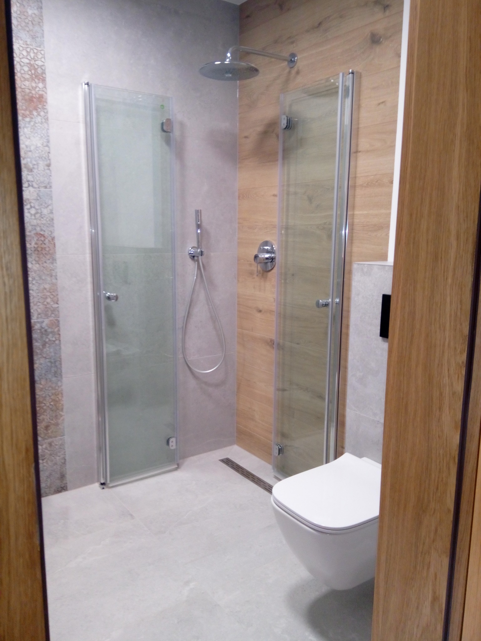 Nowoczesna łazienka z kabiną prysznicową typu walk-in, wiszącą toaletą i drewnopodobnymi panelami ściennymi. Minimalistyczny design z szarymi płytkami i chromowanymi elementami.