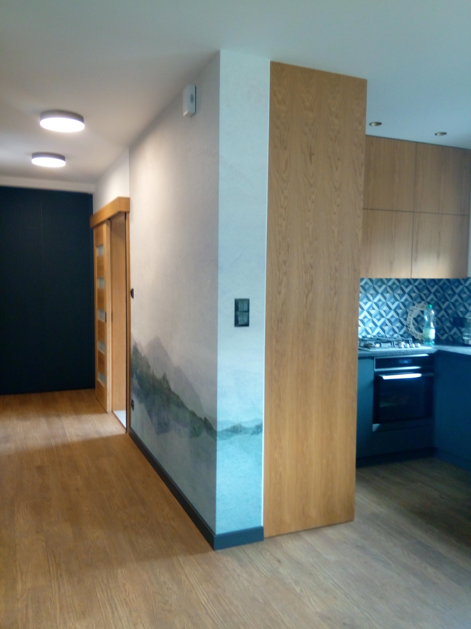 Nowoczesne wnętrze: drewniana podłoga, ściana z motywem górskim, drewniane drzwi i zabudowa kuchenna z niebieskimi akcentami. Oświetlenie sufitowe.