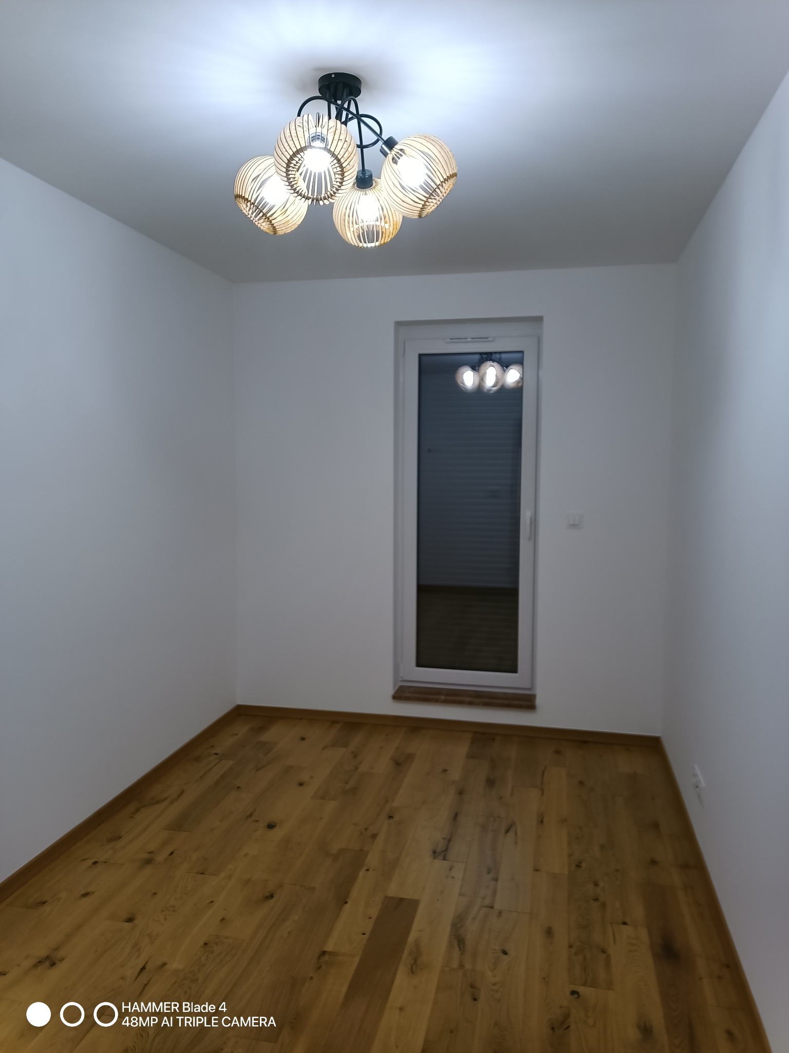 Pusty pokój z drewnianą podłogą i białymi ścianami, oświetlony nowoczesną lampą sufitową. Widoczne okno z roletami. Minimalistyczne wykończenie wnętrza.