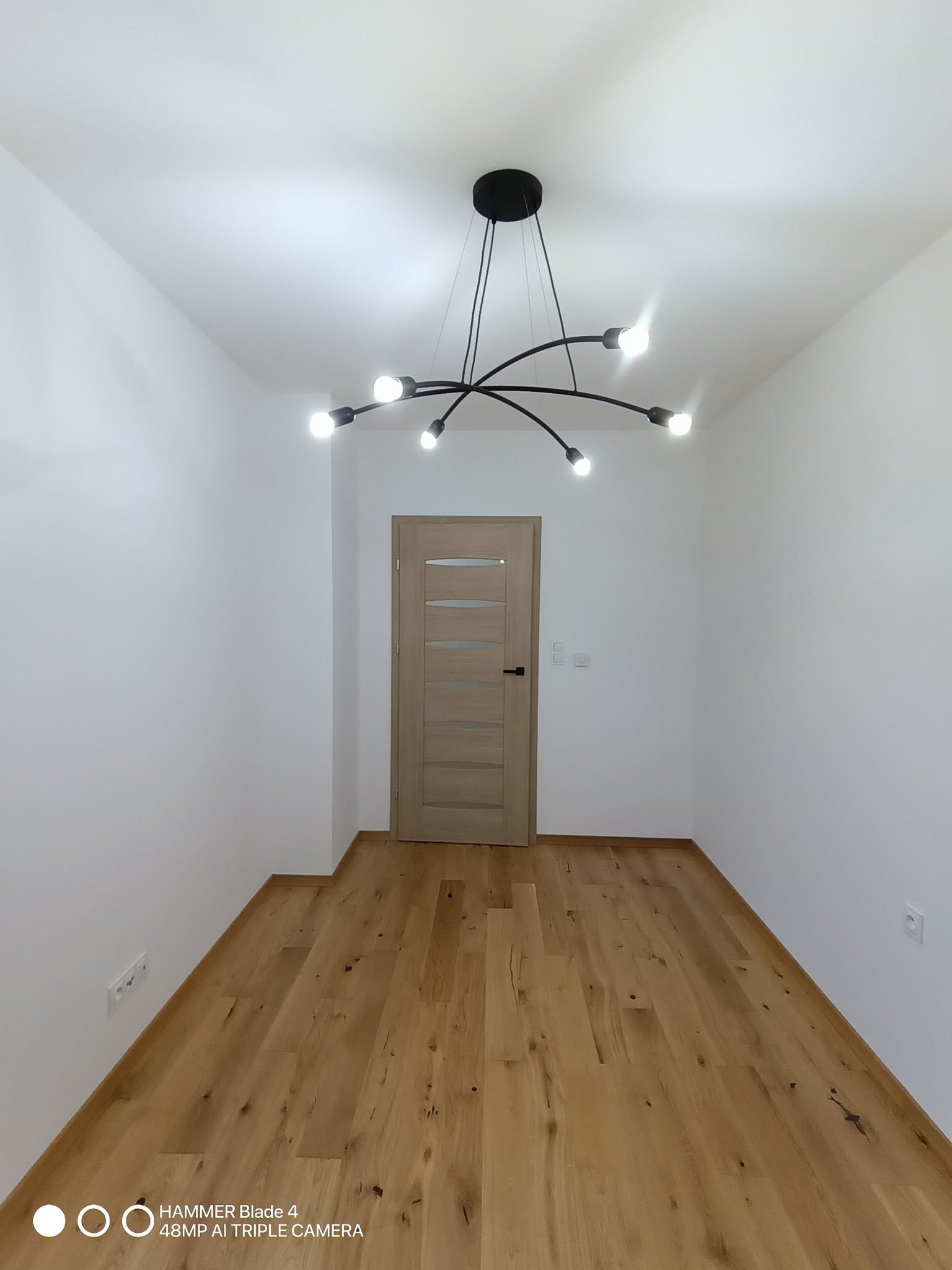 Wnętrze pokoju po remoncie: drewniana podłoga, białe ściany i nowoczesna, czarna lampa sufitowa. Widoczne gniazdka elektryczne i drzwi z przeszkleniem.