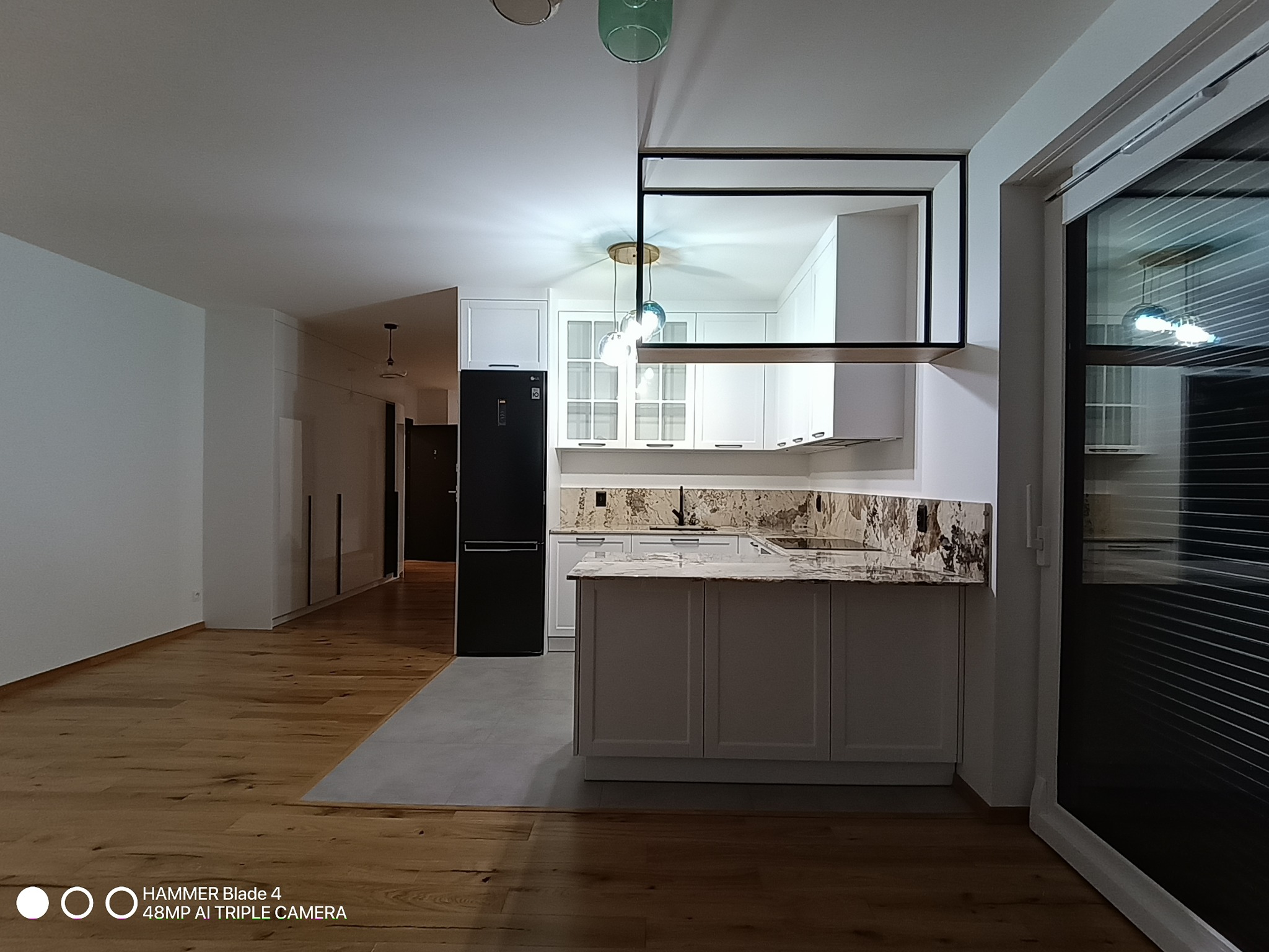 Nowoczesna, jasna kuchnia z wyspą i drewnianą podłogą. Widoczne białe szafki, czarna lodówka i marmurowy blat. Minimalistyczny design z otwartą przestrzenią.