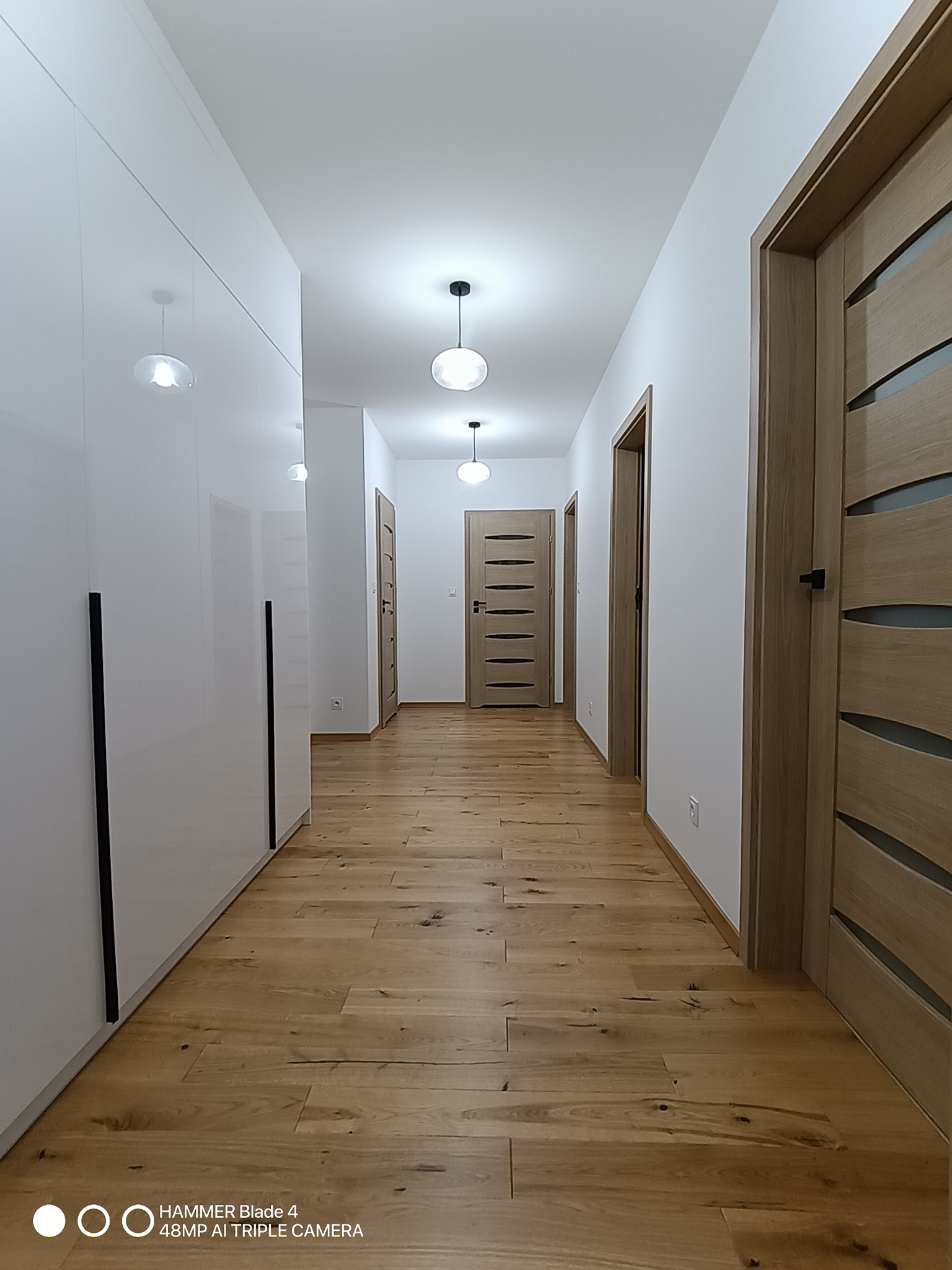 Nowoczesny korytarz z drewnianą podłogą, białymi ścianami i szafą w zabudowie. Widoczne drzwi wewnętrzne z elementami szkła i minimalistyczne oświetlenie sufitowe.