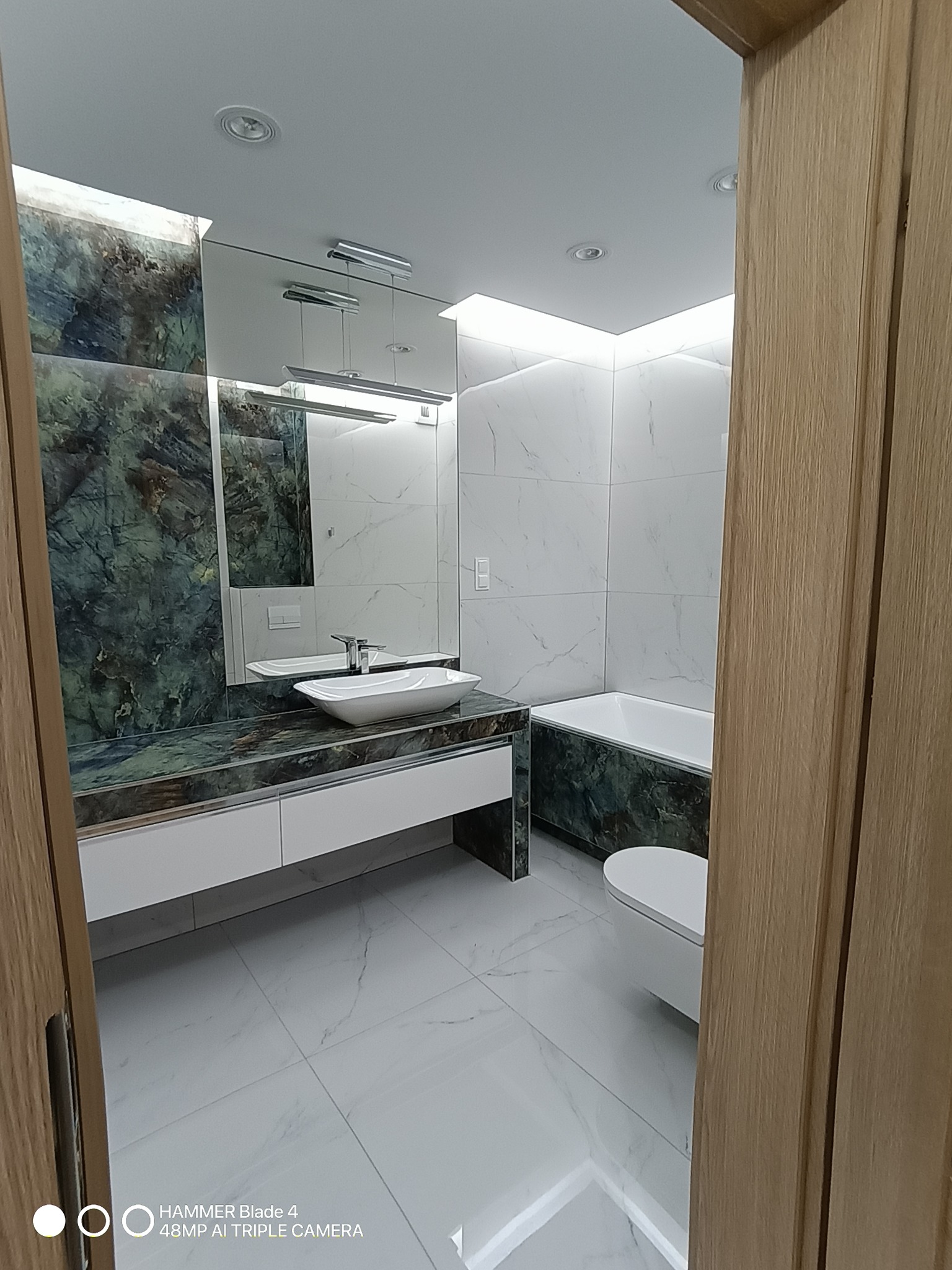 Nowoczesna łazienka z marmurowymi płytkami, umywalką na blacie z zielonego marmuru i oświetleniem LED, widziana zza uchylonych drewnianych drzwi.