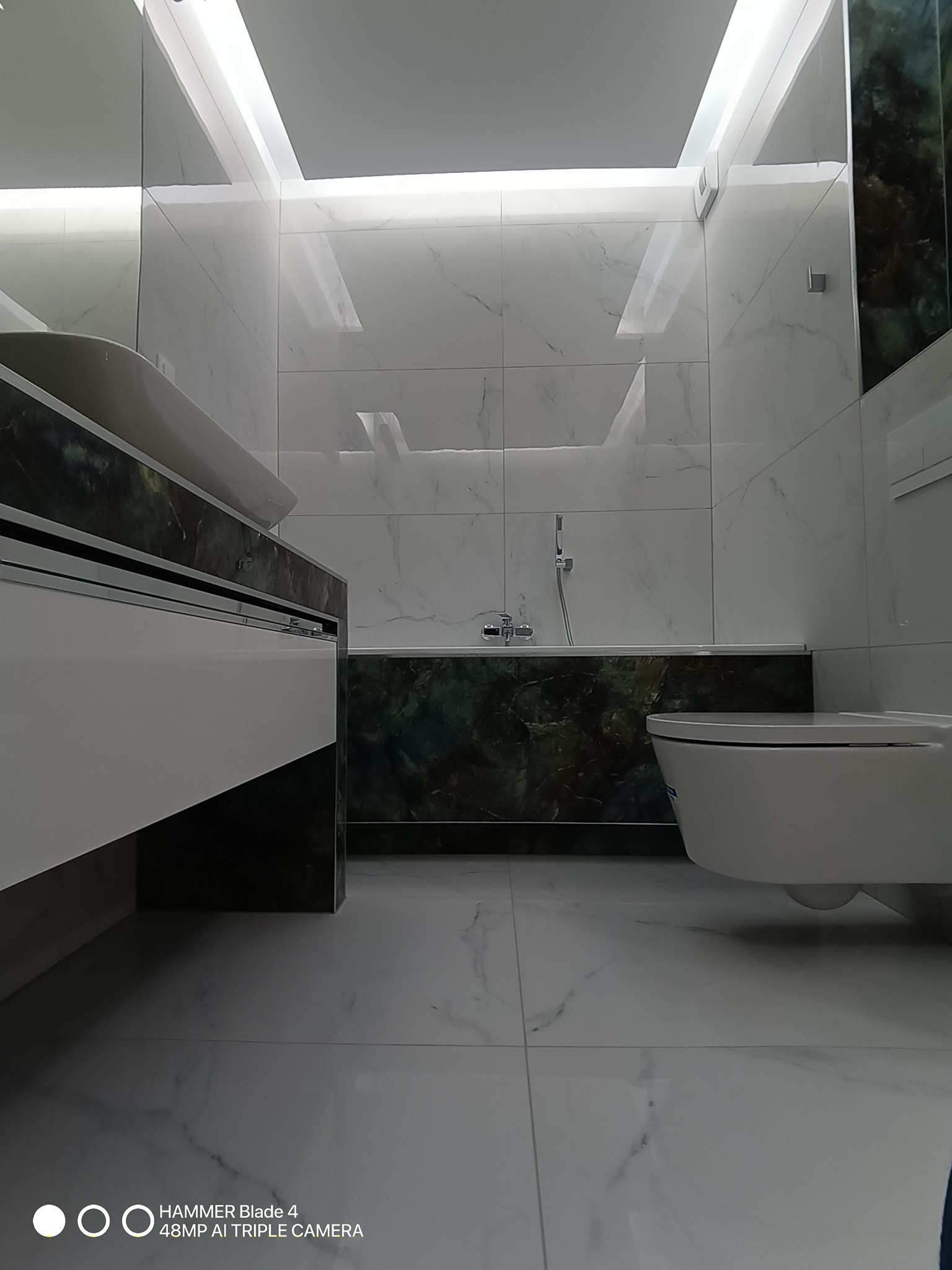 Nowoczesna łazienka z marmurowymi płytkami, podświetlanym sufitem i elementami w odcieniach zieleni. Uwagę przykuwa wanna obudowana płytkami imitującymi zielony marmur.