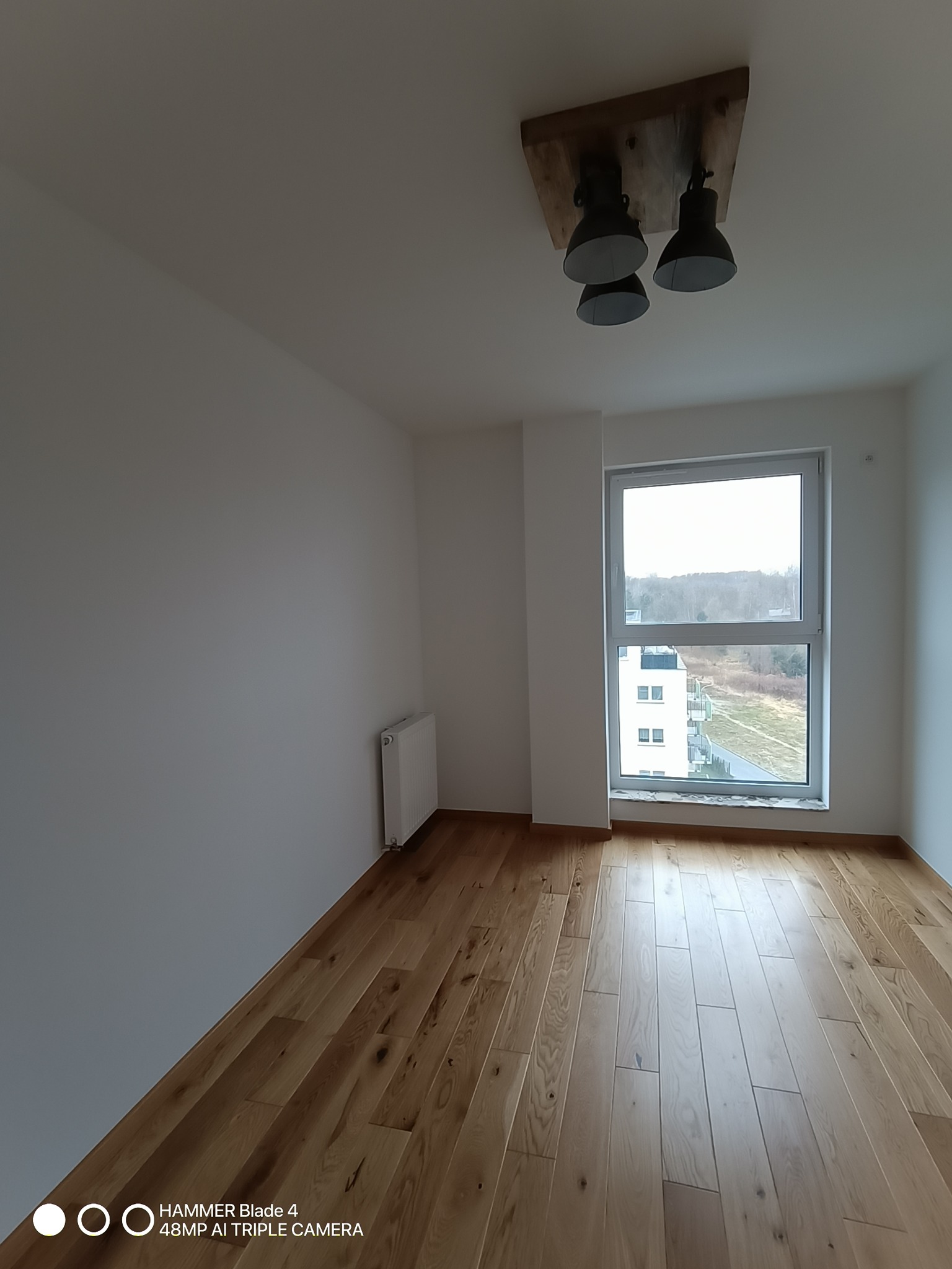 Jasne, puste wnętrze z drewnianą podłogą, białymi ścianami, grzejnikiem i oknem z widokiem na zieleń. Nietypowa, drewniana lampa sufitowa z trzema kloszami.