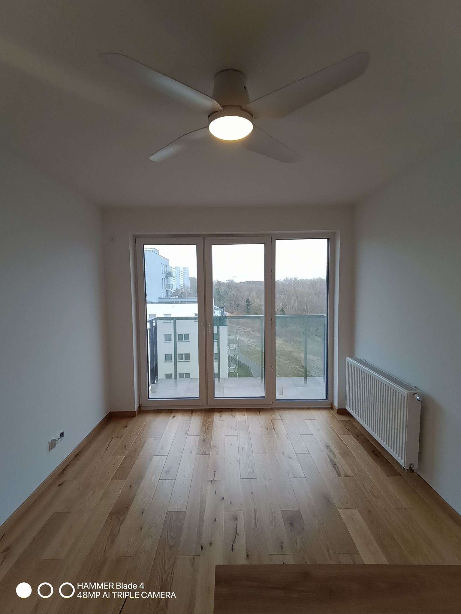 Jasne wnętrze z drewnianą podłogą, białymi ścianami, grzejnikiem i wentylatorem sufitowym. Widok z okna na budynki i drzewa. Minimalistyczny design.