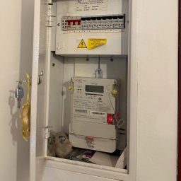 Otwarta skrzynka elektryczna z licznikiem i bezpiecznikami wewnątrz, widoczne klucze na drzwiach. Żółta naklejka ostrzegawcza o niebezpieczeństwie porażenia.