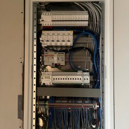 Rozdzielnica elektryczna w metalowej obudowie, z licznymi przewodami oznaczonymi etykietami, widoczne zabezpieczenia i moduły, schludne ułożenie kabli.