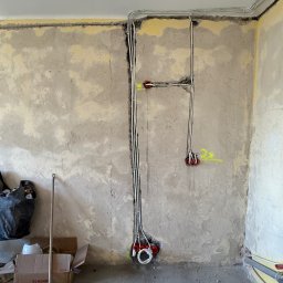Dobra Faza - Ściana w trakcie remontu z widocznymi przewodami elektrycznymi w peszlach, prowadzącymi do puszek instalacyjnych. Na podłodze karton i narzędzia.