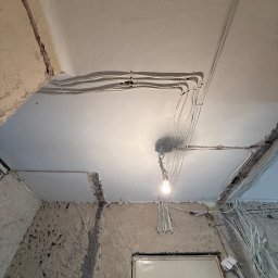 Dobra Faza - Białe przewody elektryczne ułożone na suficie w surowym wnętrzu. Widoczna żarówka i fragmenty ścian w stanie deweloperskim. Instalacja w trakcie prac wykończeniowych.