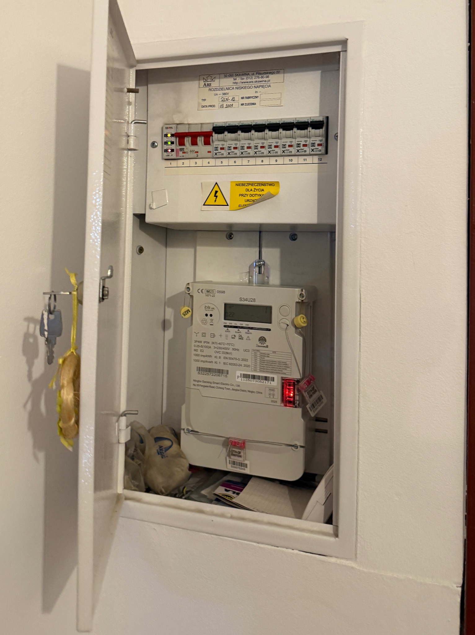 Otwarta skrzynka elektryczna z licznikiem i bezpiecznikami wewnątrz, widoczne klucze na drzwiach. Żółta naklejka ostrzegawcza o niebezpieczeństwie porażenia.