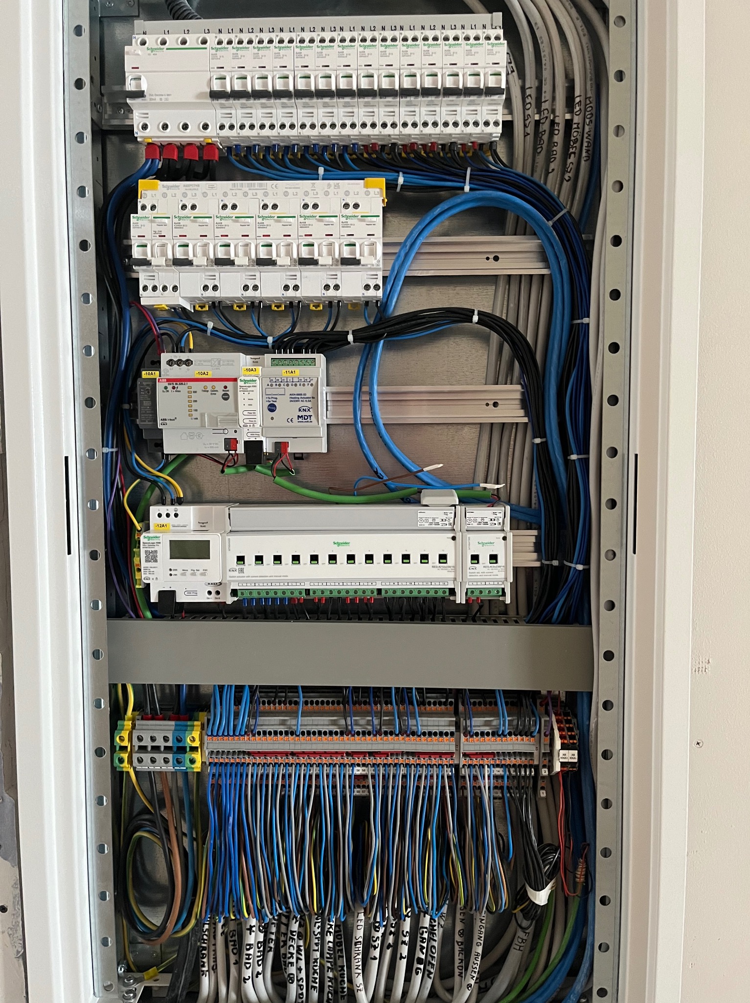 Rozdzielnia elektryczna z licznymi przewodami, zabezpieczeniami i modułami, starannie ułożona w metalowej obudowie. Widoczne oznaczenia przewodów i komponentów Schneider.