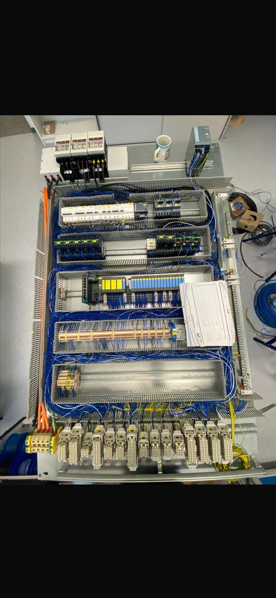 Rozbudowana szafa sterownicza z licznymi przewodami, modułami i zabezpieczeniami. Widoczne elementy automatyki przemysłowej i złącza. Na górze kubek.