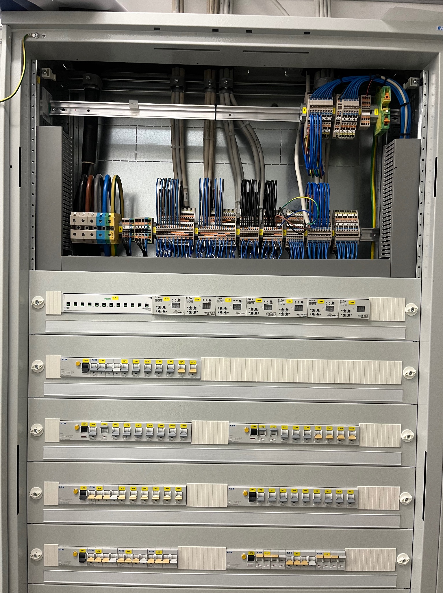 Rozdzielnica elektryczna w szafie, widoczne bezpieczniki, przewody i złącza. Profesjonalny montaż w szarej obudowie.