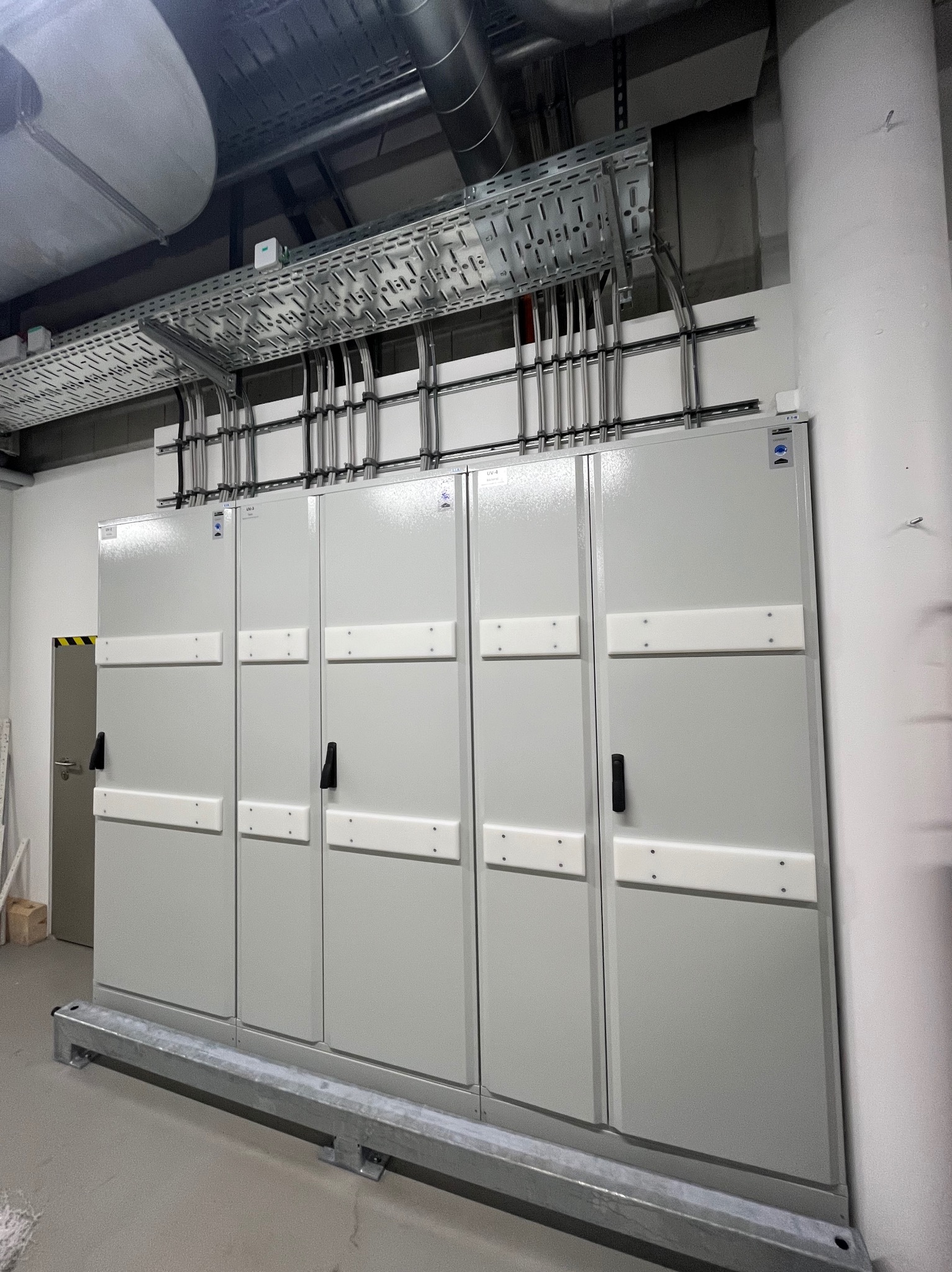 Szara rozdzielnica elektryczna z przewodami w metalowych korytach, zamontowana w pomieszczeniu technicznym. Widoczne elementy mocujące i oznaczenia na obudowie.