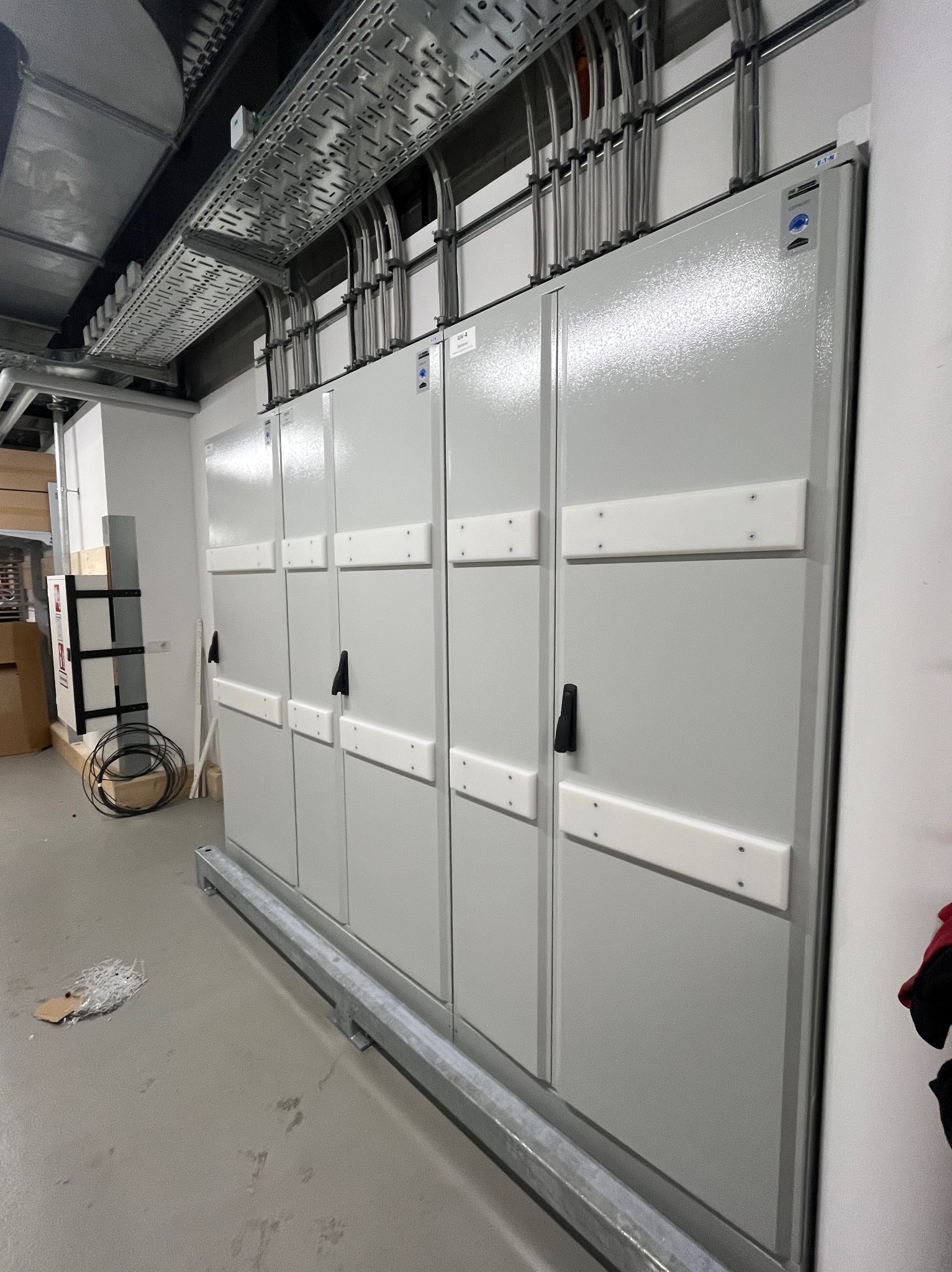 Szara rozdzielnica elektryczna z przewodami w rurach ochronnych, zamontowana w pomieszczeniu technicznym. Widoczne korytka kablowe pod sufitem i fragment podłogi.