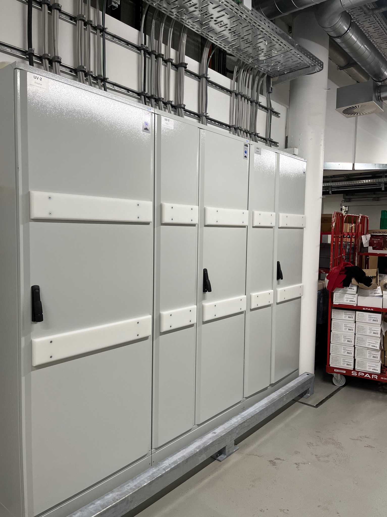 Szara rozdzielnica elektryczna z czarnymi uchwytami, widoczne kable w metalowych korytach pod sufitem, w tle wózek z kartonami.