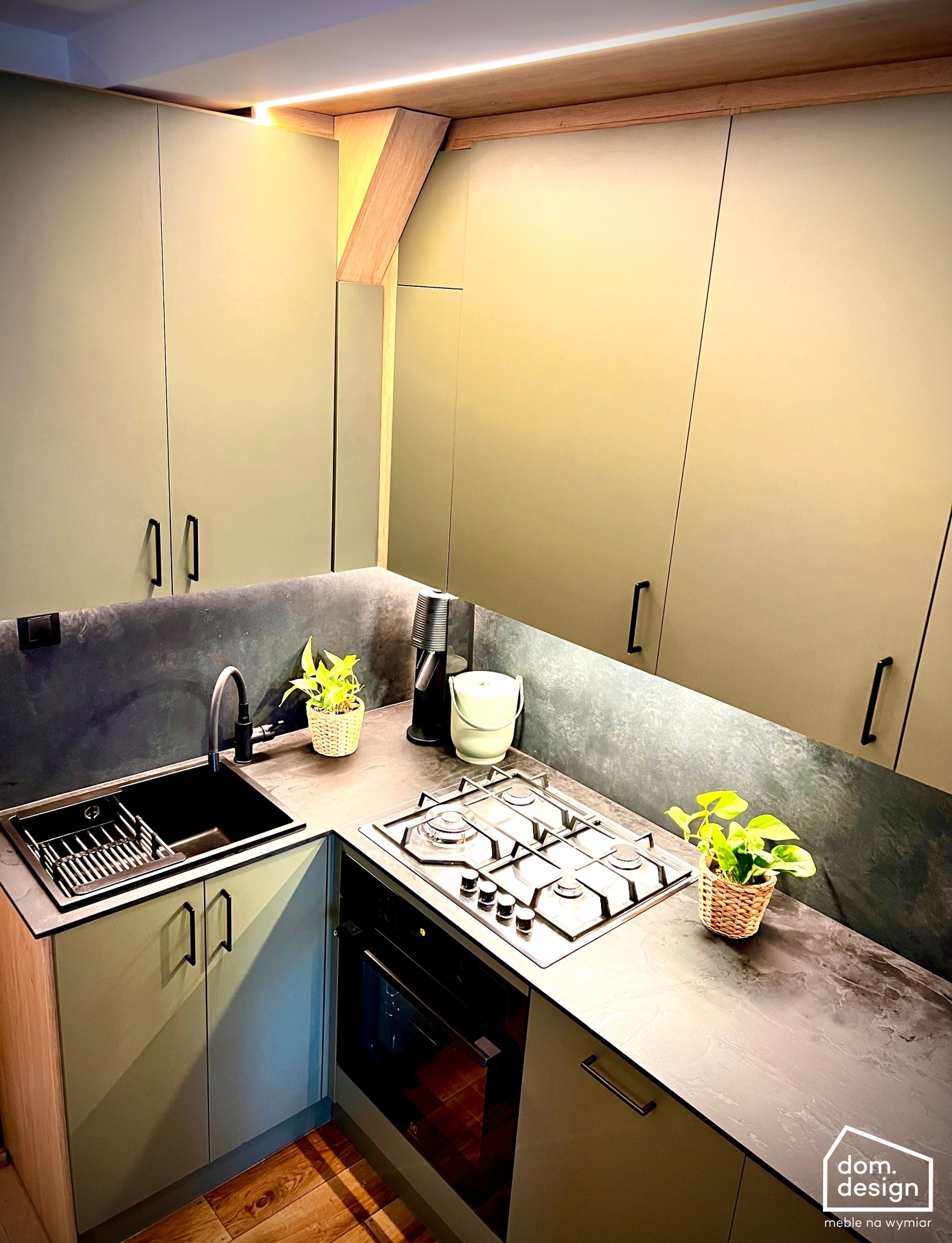 Oliwkowa kuchnia na wymiar z blatem HPL i fornirem kamiennym, oświetlona LED. Ujęcie z góry ukazuje detale wykończenia i aranżację z roślinami doniczkowymi.