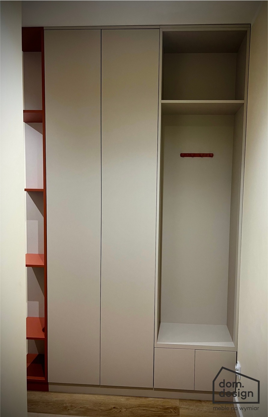 Jasna szafa wnękowa z czerwonymi akcentami: półki i wieszaki. Minimalistyczny design, zabudowa od podłogi do sufitu. Widoczne logo firmy meblarskiej.