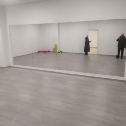 Ruslan Alyshov - Jasne wnętrze sali z zamontowanymi lustrami na ścianie. Widoczne osoby odbijające się w lustrach i podłoga z paneli. Minimalistyczny wystrój.