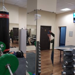 Ruslan Alyshov - Montaż lustra w siłowni: lustrzane ściany z widocznymi mocowaniami, odbijające sprzęt do ćwiczeń i osobę robiącą zdjęcie. Widoczne obciążenia i worek bokserski.