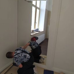 Ruslan Alyshov - Montaż dużego lustra na ścianie w trakcie pracy. Widoczne narzędzia i osoba w trakcie instalacji. Lustro odbija okno i drugą osobę.
