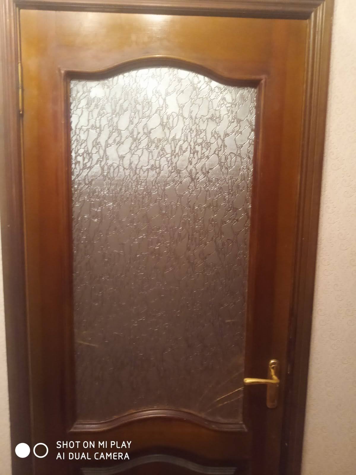 Brązowe drzwi wewnętrzne z ozdobnym szkłem i złotą klamką, widoczne na tle ściany z tapetą. Drzwi z wyraźnymi detalami ramy i wzoru szkła.