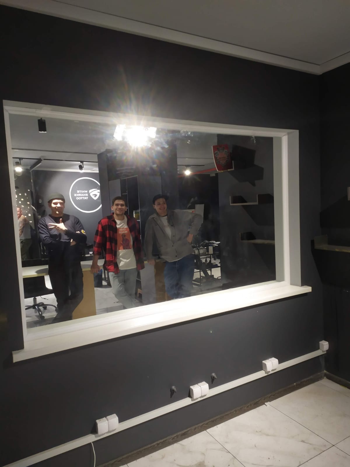 Nowe okno z trzema osobami odbijającymi się w szybie, szara ściana z białymi listwami przypodłogowymi i gniazdkami. Widok na wnętrze lokalu.