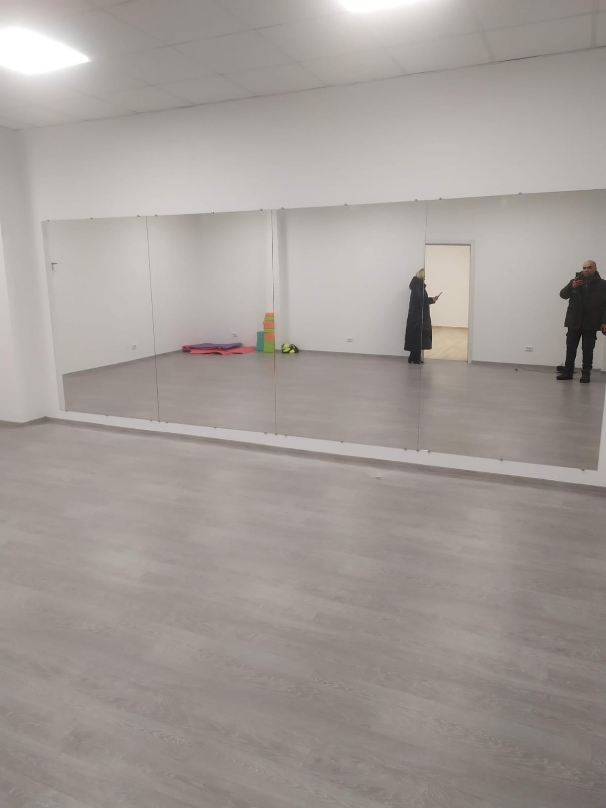 Jasne wnętrze sali z zamontowanymi lustrami na ścianie. Widoczne osoby odbijające się w lustrach i podłoga z paneli. Minimalistyczny wystrój.