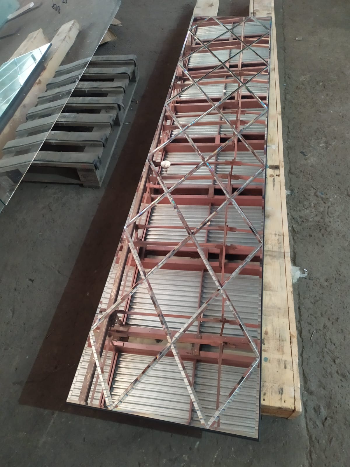 Długie lustro z geometrycznym wzorem rombów, oparte o drewnianą paletę, obok stosu szkła. Widoczne elementy konstrukcyjne i surowy beton na podłodze.