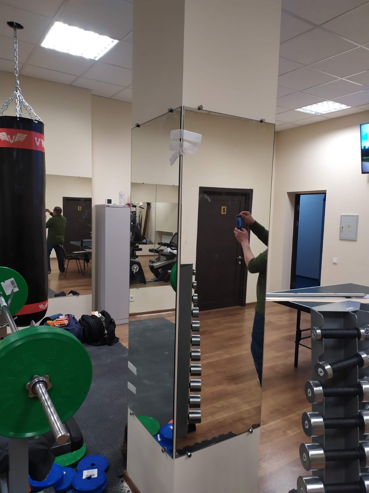Montaż lustra w siłowni: lustrzane ściany z widocznymi mocowaniami, odbijające sprzęt do ćwiczeń i osobę robiącą zdjęcie. Widoczne obciążenia i worek bokserski.