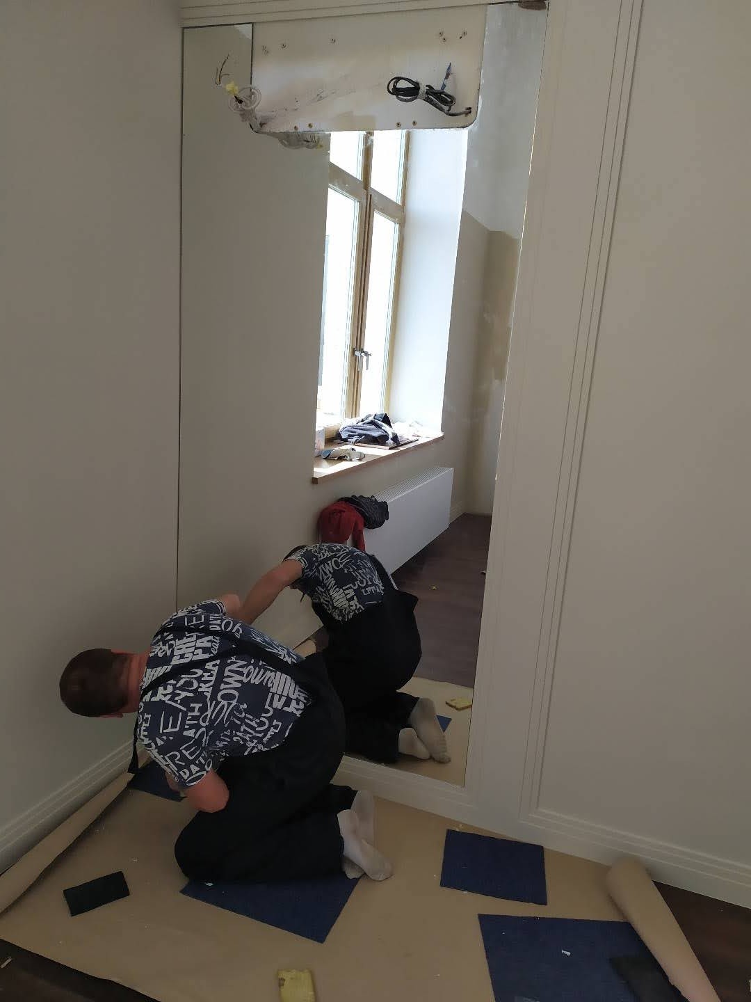 Montaż dużego lustra na ścianie w trakcie pracy. Widoczne narzędzia i osoba w trakcie instalacji. Lustro odbija okno i drugą osobę.