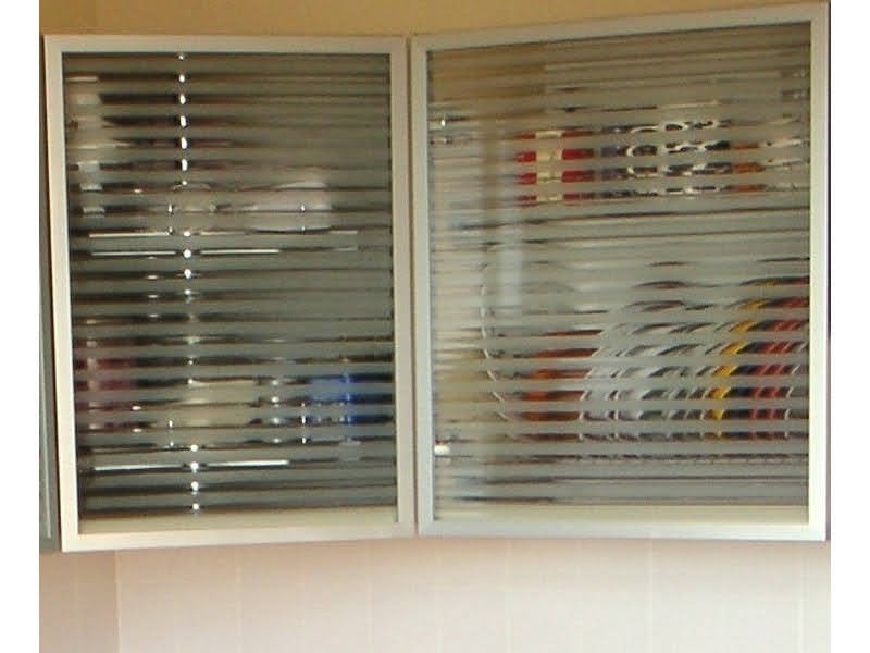 Dwa okna szafki kuchennej z poziomymi żaluzjami, częściowo zasłonięte matową folią, ukazujące zawartość szafki w tle. Jasne ramy okien.