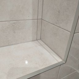 Michał Guźniczak - Narożnik wyłożony jasnymi, matowymi płytkami ceramicznymi z delikatną fakturą. Widoczne fugi i listwa wykończeniowa. Minimalistyczny design.