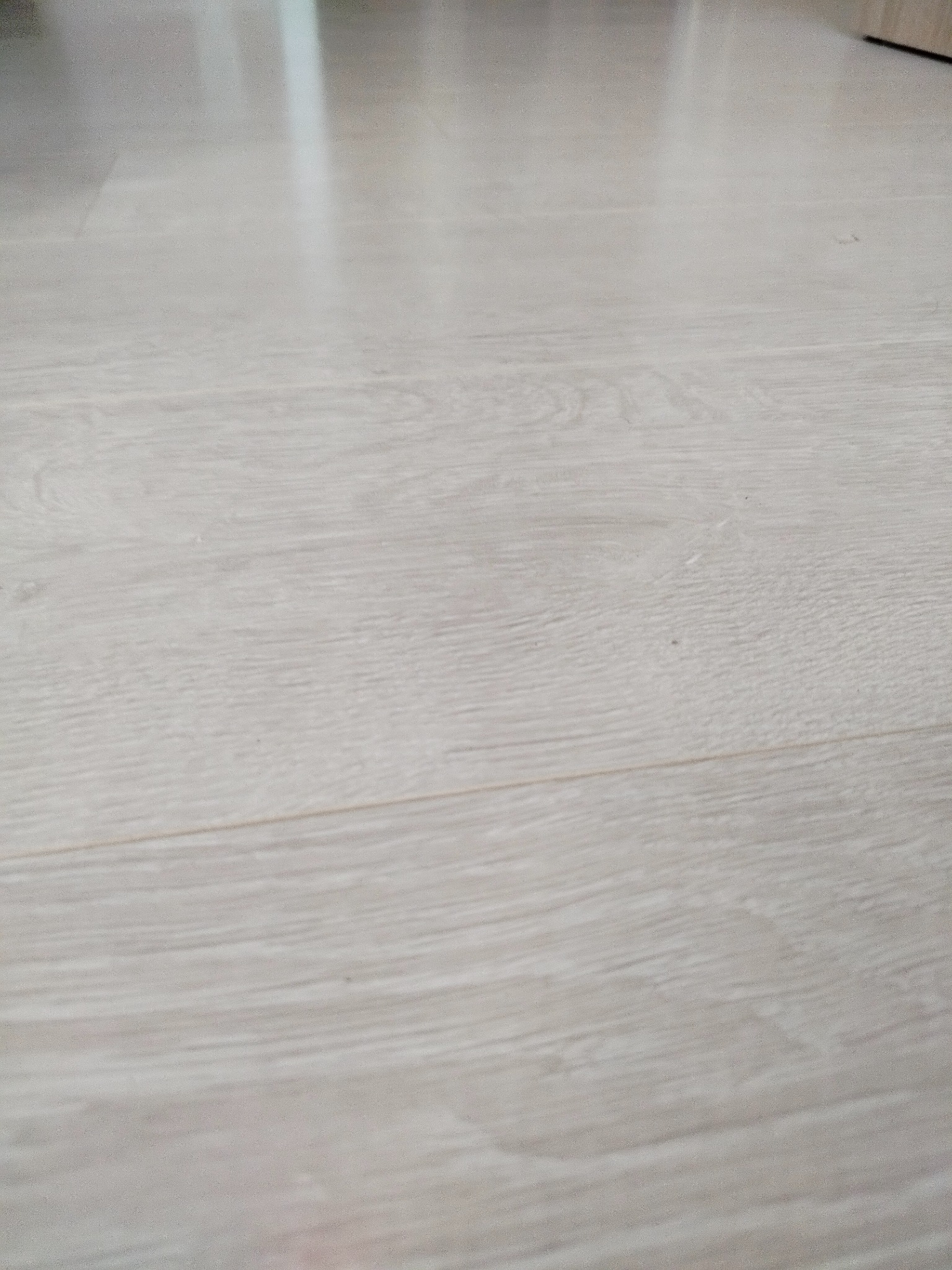 Jasna, drewnopodobna podłoga laminowana, ułożona wzdłużnie, z delikatnym połyskiem i widoczną strukturą drewna. Minimalistyczne ujęcie, skupiające się na fakturze i kolorystyce.