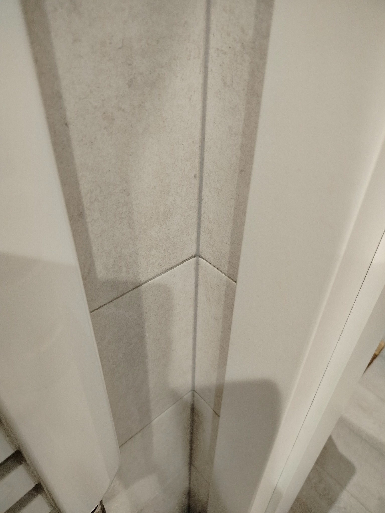 Detal wykończenia łazienki: precyzyjne połączenie płytek ceramicznych w rogu, z widoczną fugą i fragmentem białej ościeżnicy drzwiowej. Minimalistyczny design.