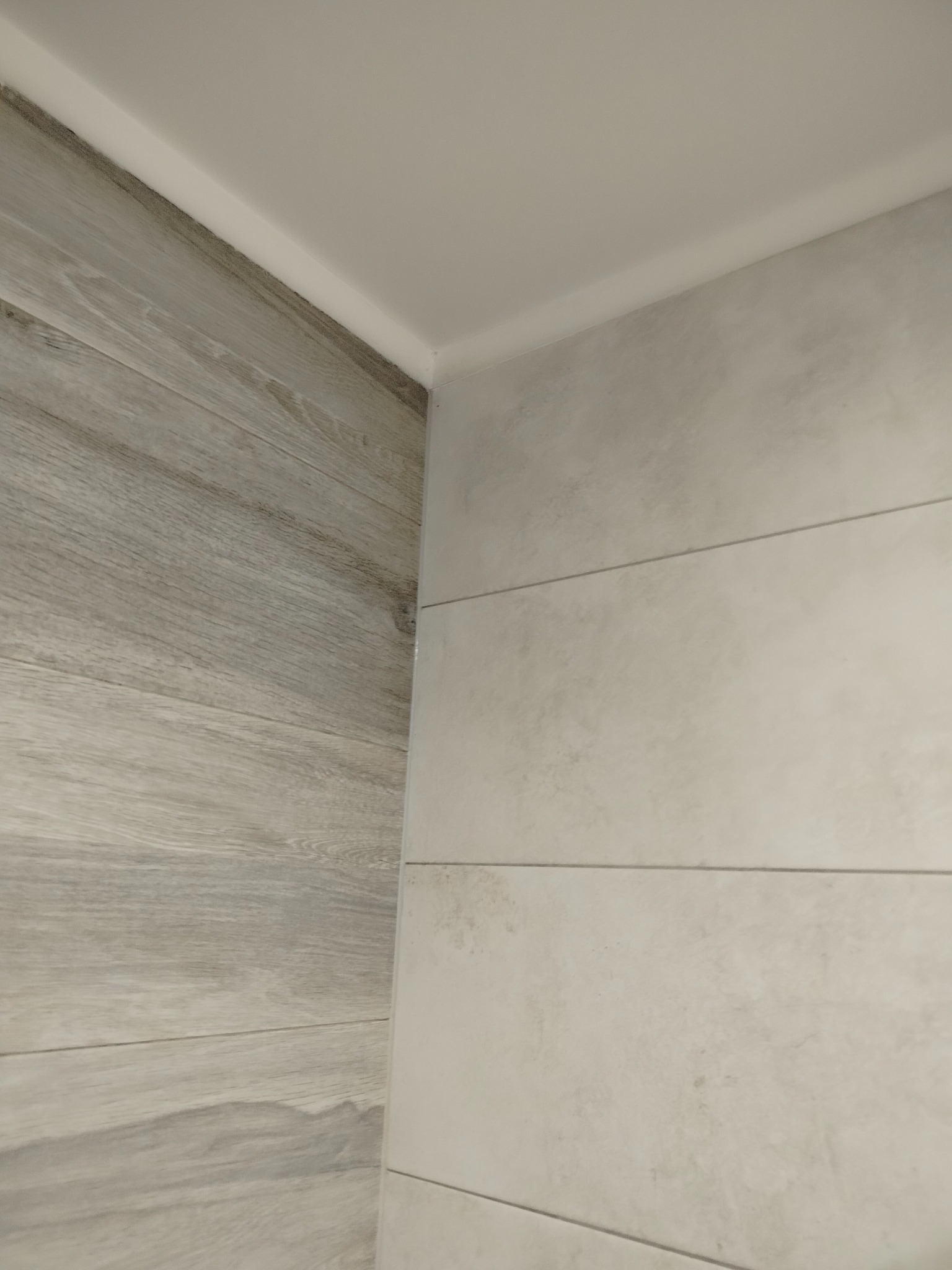 Połączenie płytek imitujących drewno i szarych, poziomych płytek w rogu pomieszczenia. Widoczna biała listwa sufitowa.