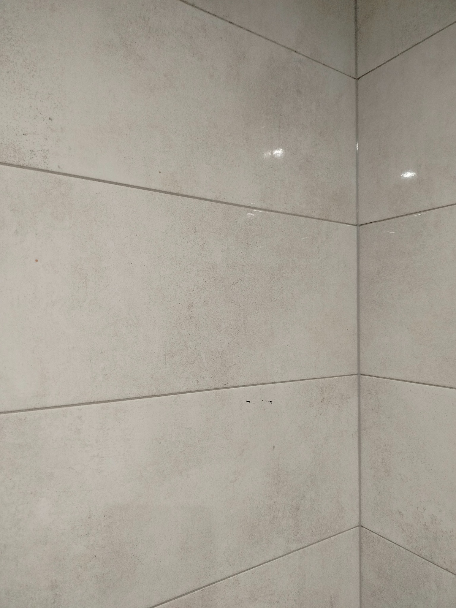 Ściana wyłożona jasnymi, matowymi płytkami ceramicznymi w formacie prostokątnym, z widocznymi fugami i delikatną strukturą powierzchni. Ujęcie z narożnika.