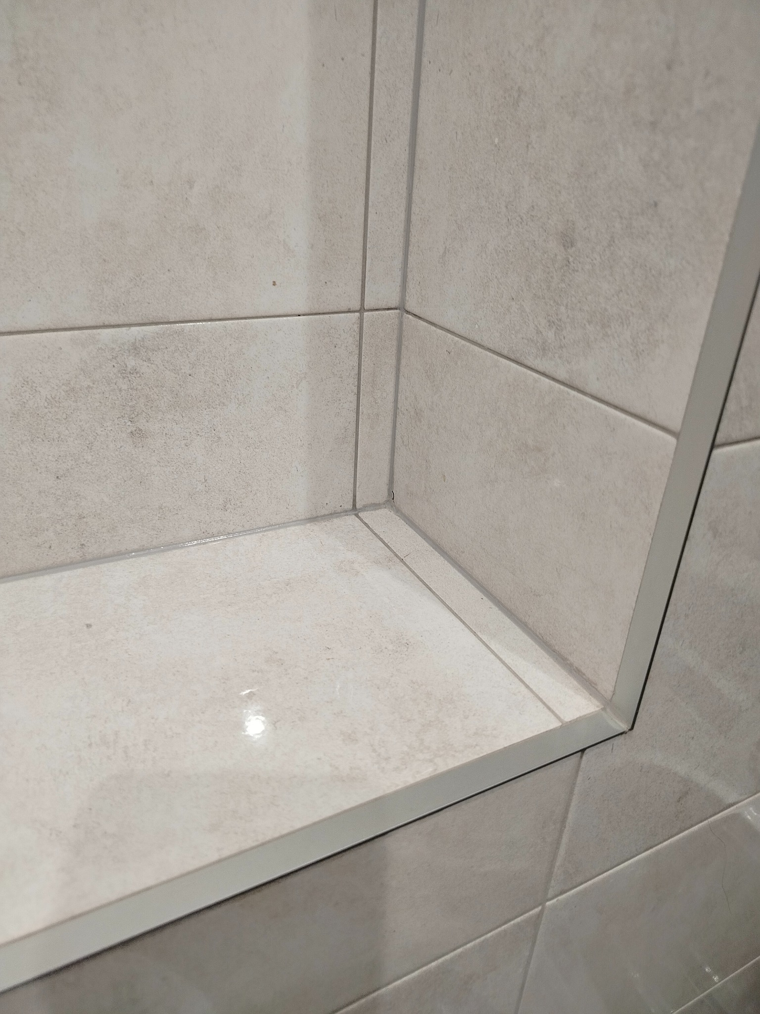 Narożnik wyłożony jasnymi, matowymi płytkami ceramicznymi z delikatną fakturą. Widoczne fugi i listwa wykończeniowa. Minimalistyczny design.