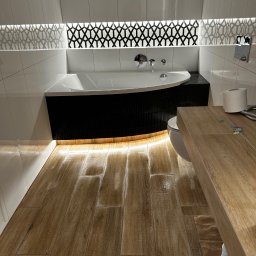 Kamil Wirkus - Nowoczesna łazienka z drewnopodobną podłogą, białą wanną obudowaną czarną mozaiką i podświetleniem LED. Geometryczny wzór nad wanną.