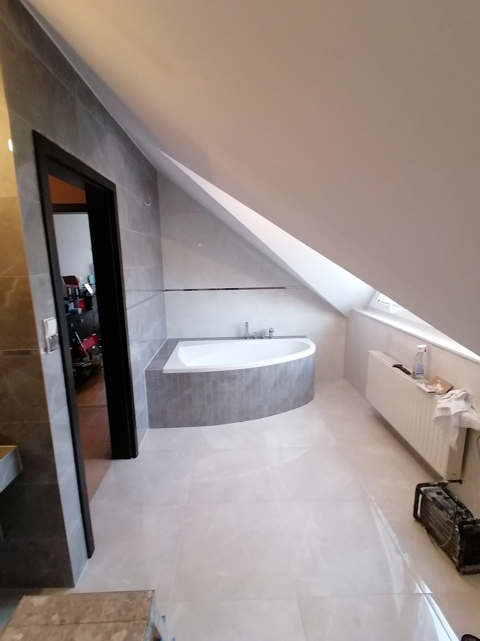 Wykończona łazienka na poddaszu ze skośnym sufitem. Szare płytki na ścianach i podłodze, wanna obudowana płytkami. Widoczny grzejnik i narzędzia.