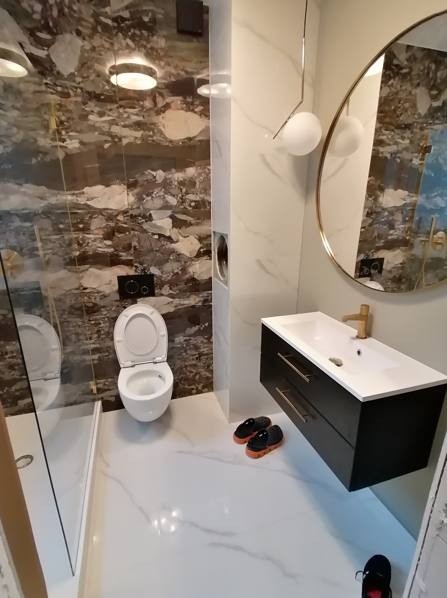 Nowoczesna łazienka z marmurową ścianą, wiszącą szafką z umywalką, okrągłym lustrem w złotej ramie i szklaną kabiną prysznicową. Minimalistyczny design z akcentami złota.