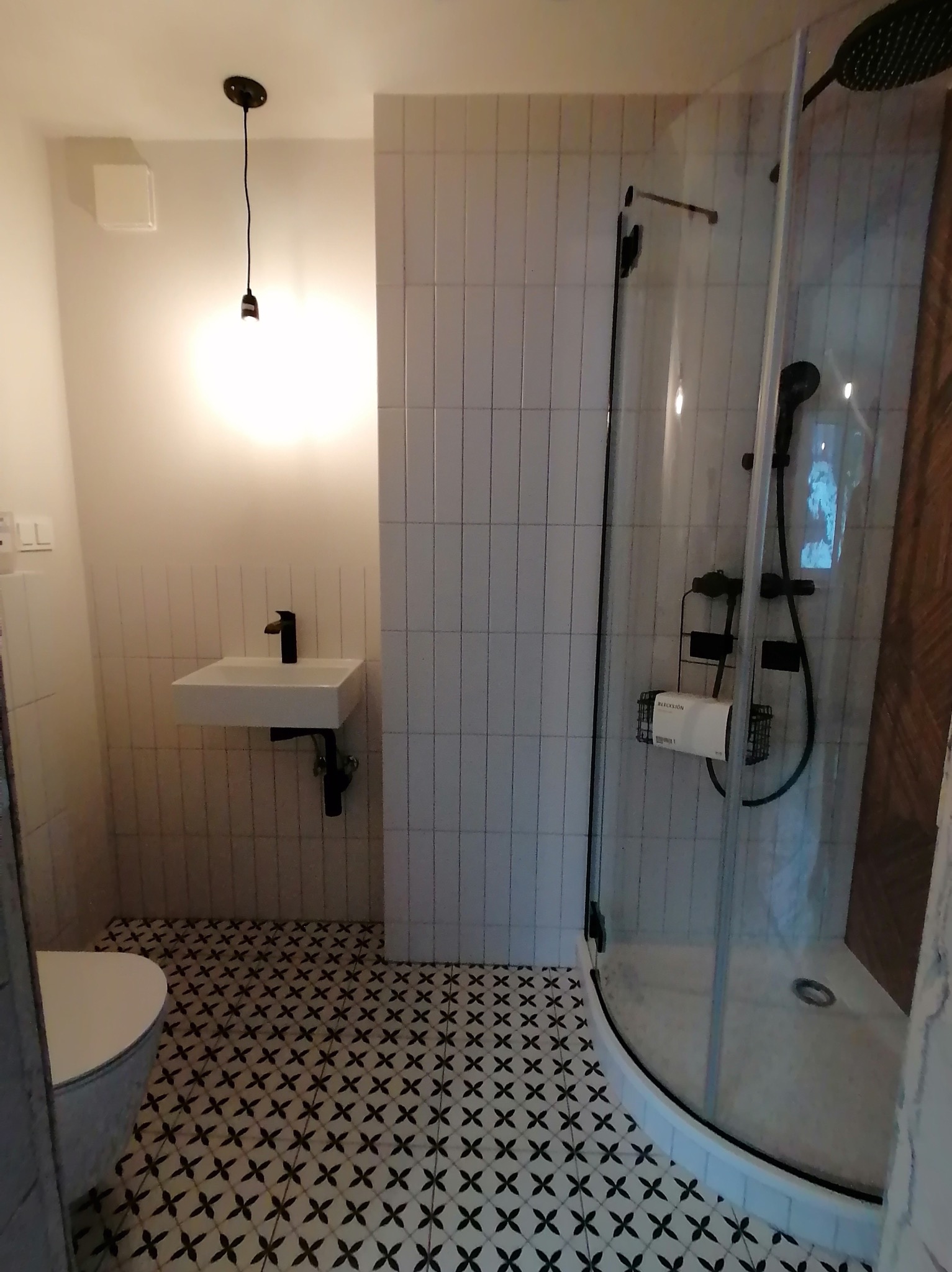 Nowoczesna łazienka z kabiną prysznicową typu walk-in, białą umywalką i czarną armaturą. Podłoga w czarno-białe wzory. Minimalistyczna lampa wisząca.