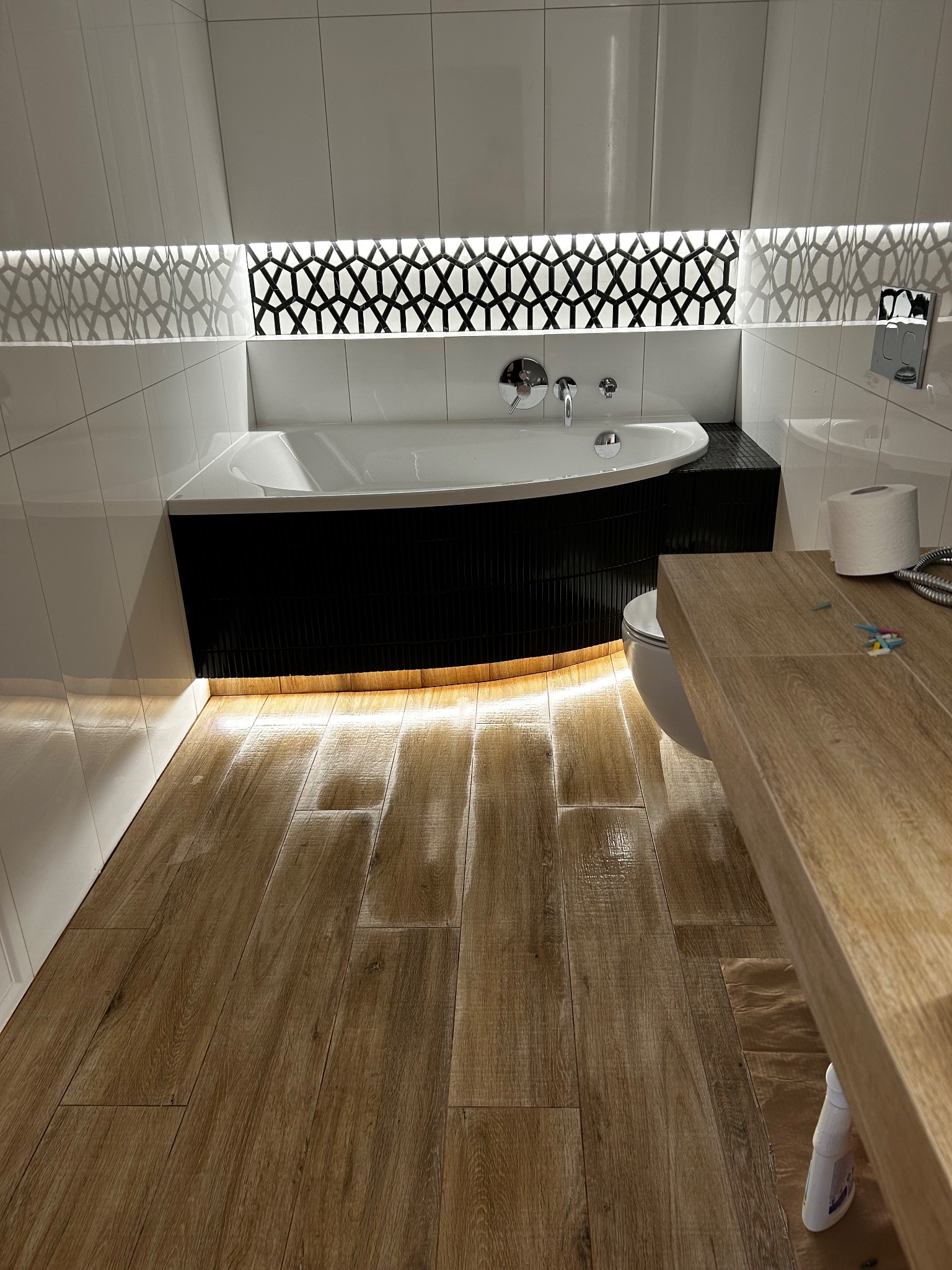 Nowoczesna łazienka z drewnopodobną podłogą, białą wanną obudowaną czarną mozaiką i podświetleniem LED. Geometryczny wzór nad wanną.