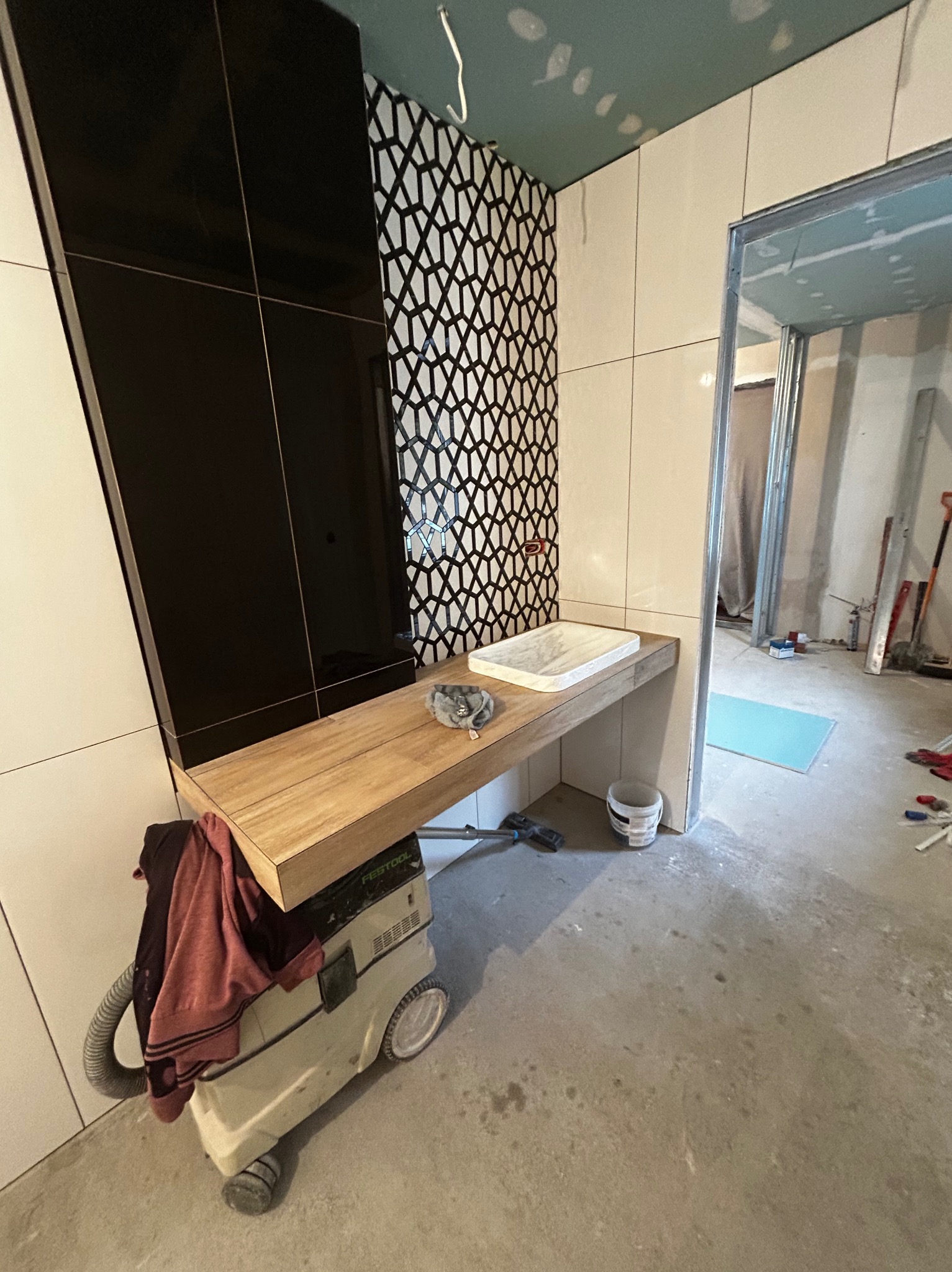 Łazienka w trakcie wykończenia: czarno-białe płytki, drewniany blat z umywalką, odkurzacz Festool. Widoczny fragment surowego pomieszczenia.